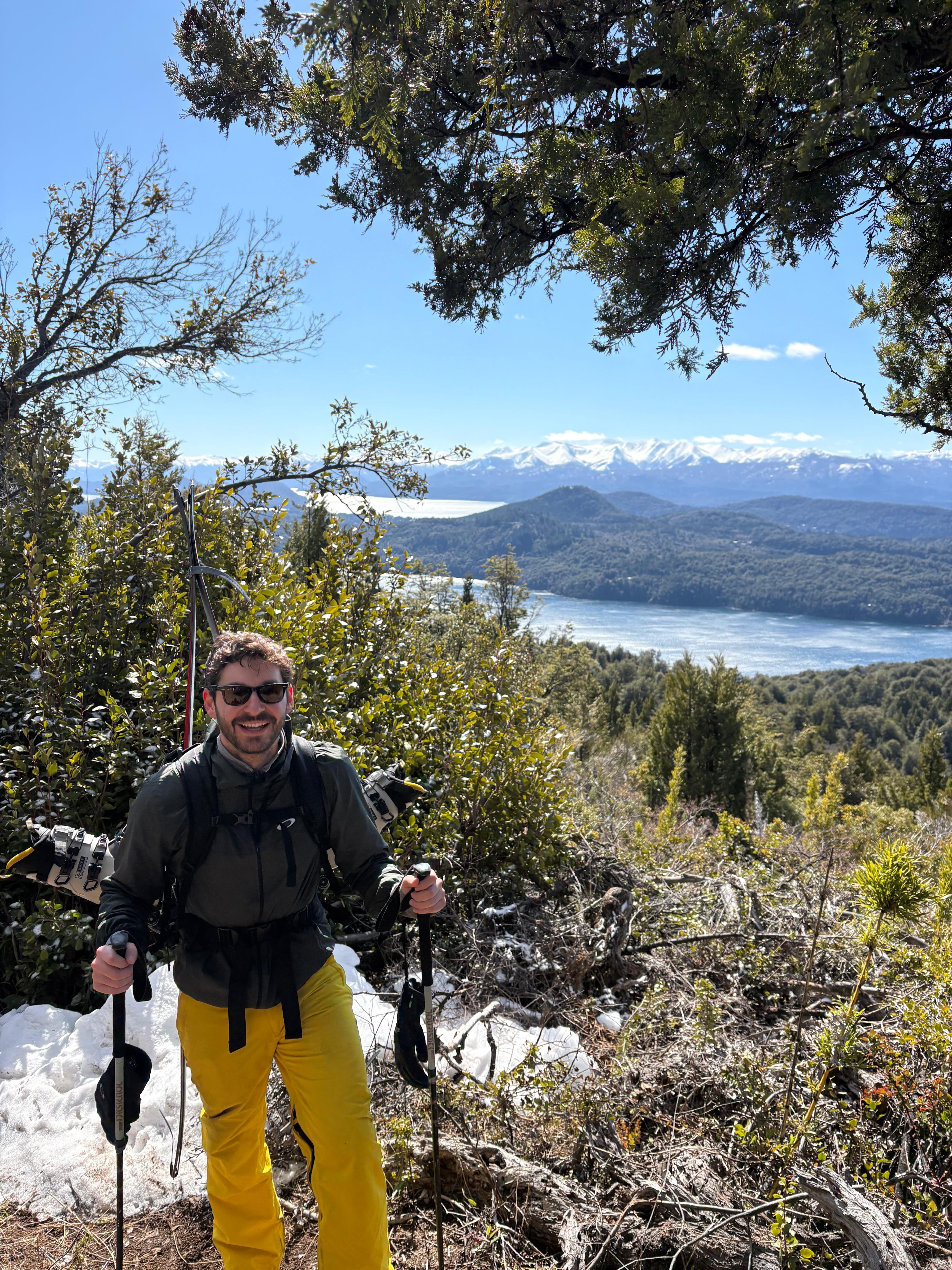 Gonzalo on a mountain trail with skis strapped to his pack, a lake and snowy peaks behind him